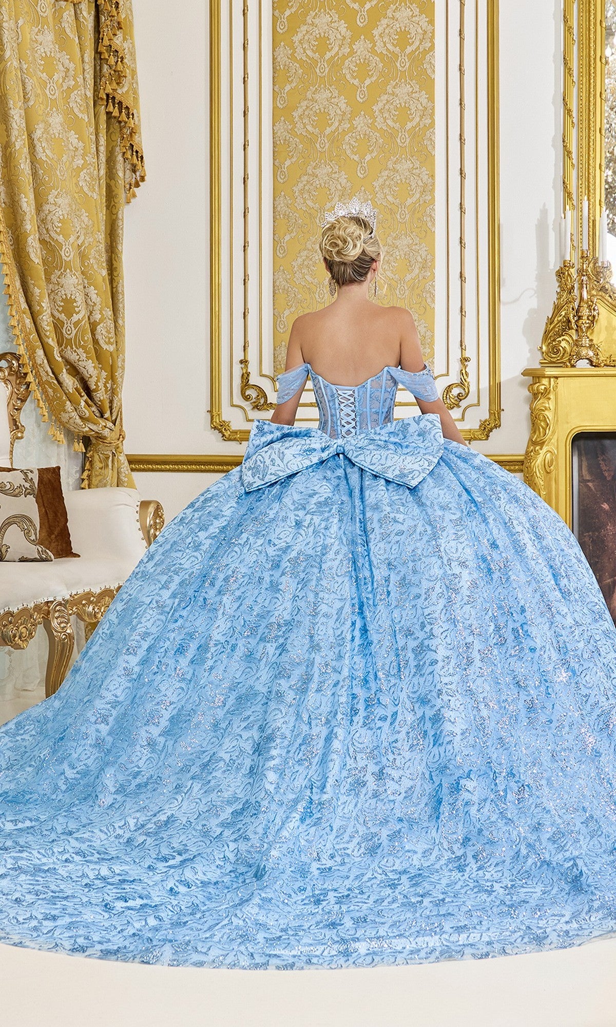 A woman in the Quinceanera Dress 5095 by Dancing Queen, featuring a blue corset bodice and large bow, stands facing away in an opulent gold-decorated room.